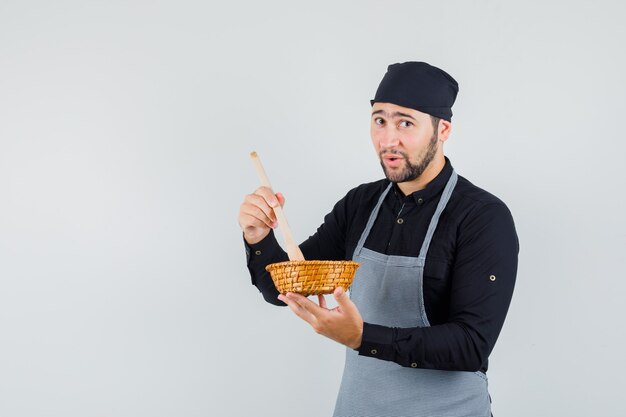 Cocinero de sexo masculino en camisa, delantal mezclando comida con cuchara de madera y luciendo linda vista frontal
