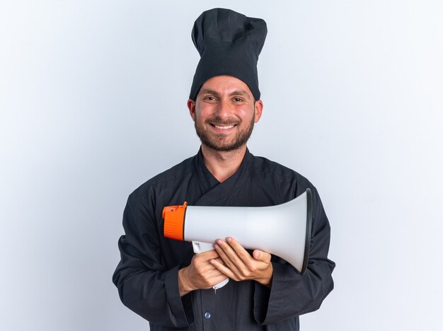 Cocinero de sexo masculino caucásico joven alegre en uniforme del cocinero y la tapa que sostiene el altavoz que mira la cámara aislada en la pared blanca