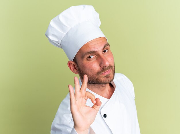 Cocinero de sexo masculino caucásico joven confiado en uniforme del cocinero y la tapa que se coloca en la vista de perfil mirando a la cámara haciendo el signo de ok aislado en la pared verde oliva