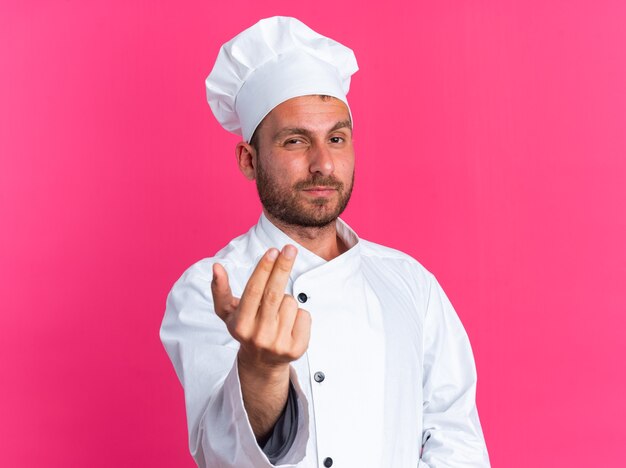 Cocinero de sexo masculino caucásico joven confiado en uniforme del cocinero y la tapa que mira la cámara haciendo gesto de venir aquí aislado en la pared rosada