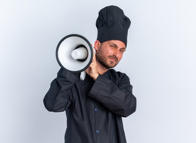 Cocinero de sexo masculino caucásico joven confiado en uniforme del cocinero y la tapa que sostiene el altavoz que mira la cámara aislada en la pared blanca con el espacio de la copia