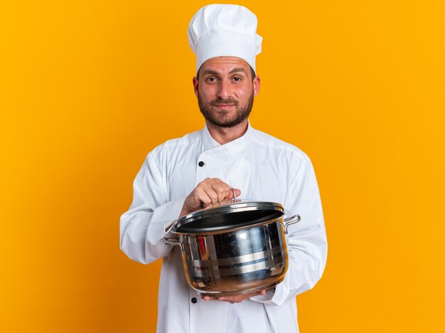 Cocinero de sexo masculino caucásico joven confiado en uniforme del cocinero y la tapa que sostiene la olla agarrando la tapa de la olla