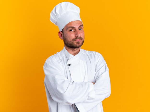 Cocinero de sexo masculino caucásico joven serio en uniforme del cocinero y la tapa que se coloca con la postura cerrada que mira la cámara aislada en la pared naranja