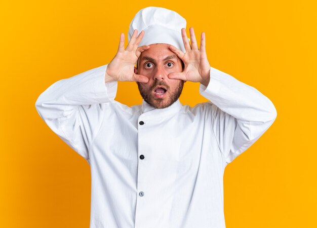 Cocinero de sexo masculino caucásico joven sorprendido en uniforme del cocinero y la tapa que mira la cámara que hace los ojos grandes aislados en la pared naranja