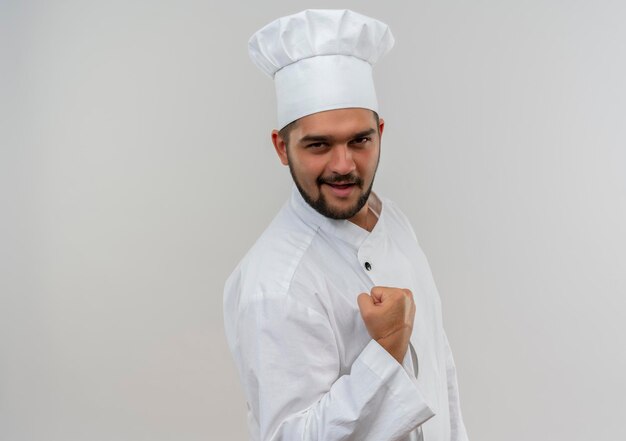 Cocinero de sexo masculino joven alegre en uniforme del cocinero que aprieta el puño aislado en la pared blanca con el espacio de la copia