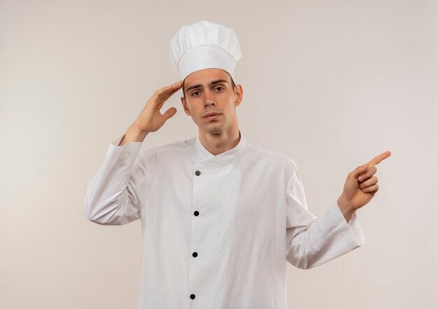 Cocinero de sexo masculino joven cansado vistiendo uniforme del cocinero que pone la mano en la frente señala el dedo al lado con el espacio de la copia