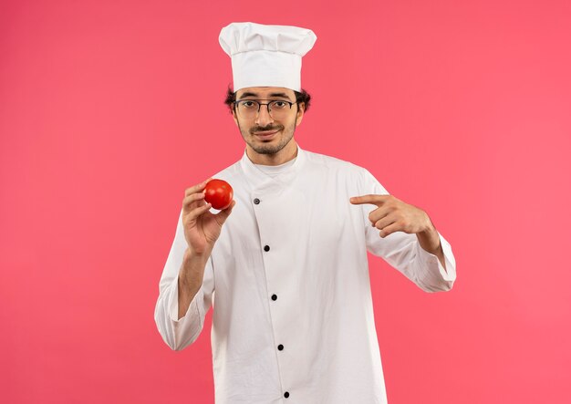 Cocinero de sexo masculino joven complacido con uniforme de chef y gafas sosteniendo y apunta a tomate aislado en la pared rosa