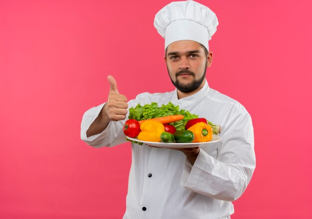 Cocinero de sexo masculino joven complacido en uniforme de chef sosteniendo un plato de verduras y mostrando el pulgar hacia arriba aislado en el espacio rosa