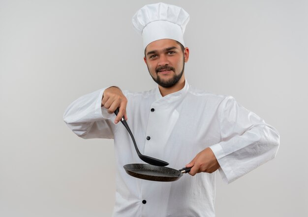 Cocinero de sexo masculino joven complacido en uniforme del cocinero que sostiene la cuchara ranurada y la sartén aislada en el espacio en blanco