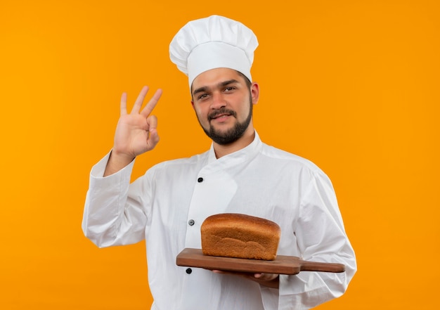 Cocinero de sexo masculino joven complacido en uniforme del cocinero que sostiene la tabla de cortar con el pan en él y que hace el signo bien aislado en el espacio naranja