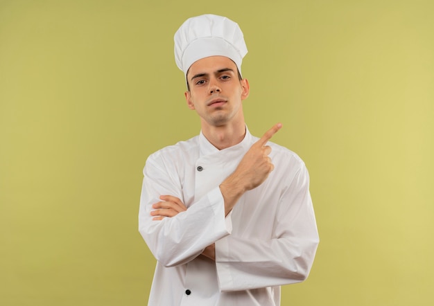 Cocinero de sexo masculino joven confiado que lleva uniforme del cocinero que cruza las manos señala el dedo al lado en la pared verde aislada con espacio de la copia