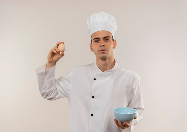 Cocinero de sexo masculino joven confiado que lleva el uniforme del cocinero que sostiene el cuenco y el huevo en la pared blanca aislada