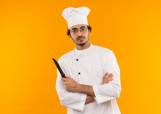Cocinero de sexo masculino joven confiado con uniforme de chef y gafas cruzando las manos y sosteniendo el cuchillo