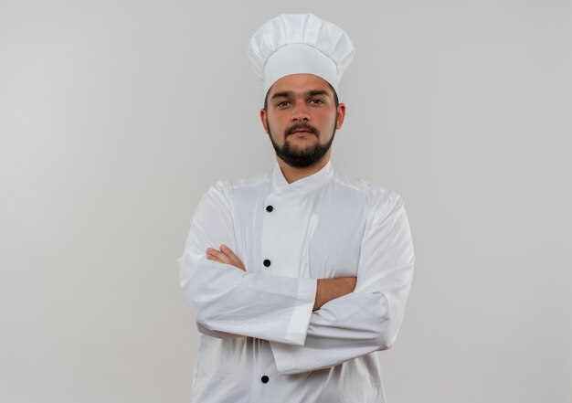 Cocinero de sexo masculino joven confiado en el uniforme del cocinero que se coloca con la postura cerrada que mira aislada en la pared blanca con el espacio de la copia