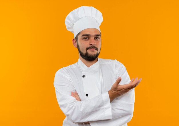 Cocinero de sexo masculino joven confiado en el uniforme del cocinero que se coloca con la postura cerrada y que muestra la mano vacía aislada en la pared naranja con el espacio de la copia