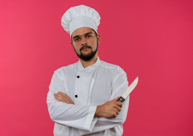 Cocinero de sexo masculino joven confiado en el uniforme del cocinero que se coloca con la postura cerrada y que sostiene el cuchillo aislado en la pared rosada