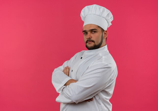 Cocinero de sexo masculino joven confiado en uniforme del cocinero que se coloca en la vista de perfil con la postura cerrada aislada en la pared rosada con el espacio de la copia