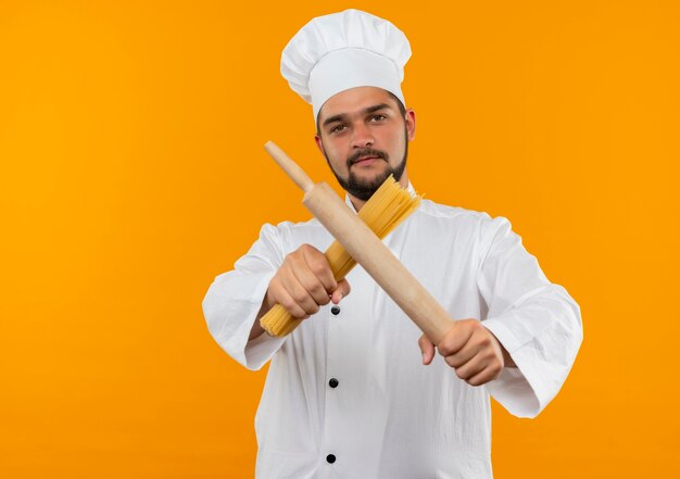 Cocinero de sexo masculino joven confiado en uniforme del cocinero que estira el rodillo y la pasta de espagueti hacia aislados en la pared naranja con espacio de copia