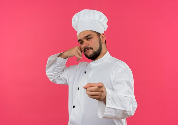 Cocinero de sexo masculino joven confiado en uniforme del cocinero que hace el gesto de la llamada y que señala aislado en la pared rosada con espacio de la copia