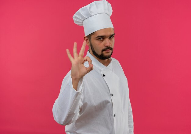Cocinero de sexo masculino joven confiado en uniforme del cocinero que hace la muestra aceptable aislada en la pared rosada con el espacio de la copia