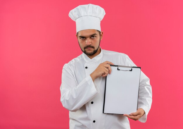 Cocinero de sexo masculino joven confiado en uniforme del cocinero que muestra el portapapeles aislado en la pared rosa con espacio de copia