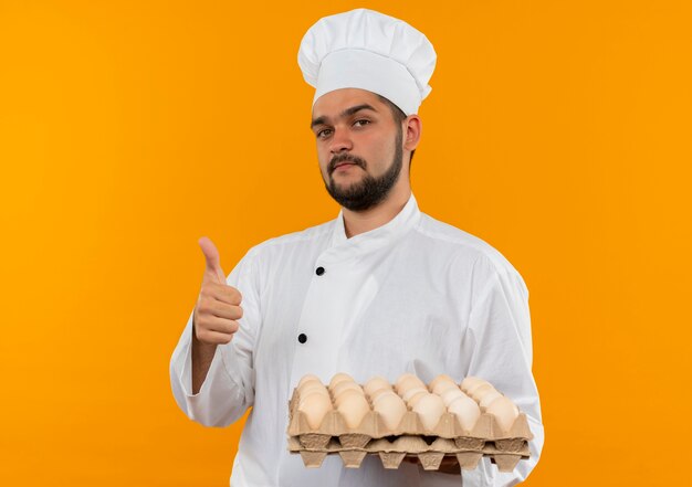 Cocinero de sexo masculino joven confiado en uniforme del cocinero que sostiene el cartón de huevos y que muestra el pulgar hacia arriba aislado en la pared naranja