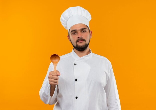 Cocinero de sexo masculino joven confiado en el uniforme del cocinero que sostiene la cuchara aislada en la pared anaranjada con espacio de copia