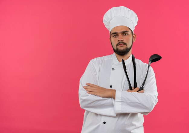 Cocinero de sexo masculino joven confiado en uniforme del cocinero que sostiene la cuchara y las pinzas aisladas en la pared rosada con espacio de la copia