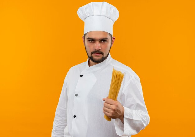 Cocinero de sexo masculino joven confiado en uniforme del cocinero que sostiene la pasta de espagueti aislada en la pared naranja con espacio de copia