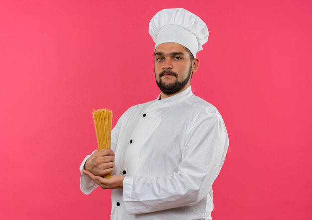 Cocinero de sexo masculino joven confiado en uniforme del cocinero que sostiene la pasta de espagueti aislada en la pared rosada con el espacio de la copia