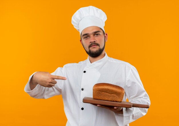 Cocinero de sexo masculino joven confiado en uniforme del cocinero que sostiene y que señala a la tabla de cortar con el pan en él aislado en la pared naranja