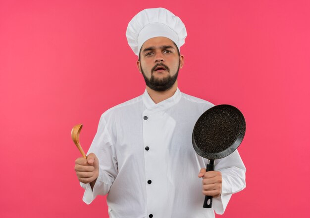 Cocinero de sexo masculino joven confundido en uniforme del cocinero que sostiene la sartén y la cuchara aislada en la pared rosada