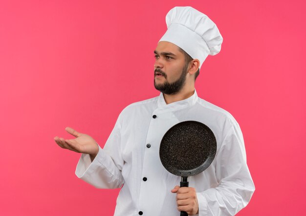 Cocinero de sexo masculino joven confundido en uniforme del cocinero que sostiene la sartén y que muestra la mano vacía que mira el lado aislado en la pared rosada