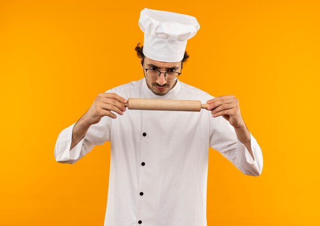 Cocinero de sexo masculino joven confundido vistiendo uniforme de chef y gafas sosteniendo y mirando el rodillo