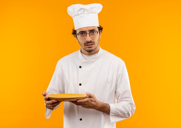 Cocinero de sexo masculino joven confundido vistiendo uniforme del cocinero y los vidrios que sostienen la placa