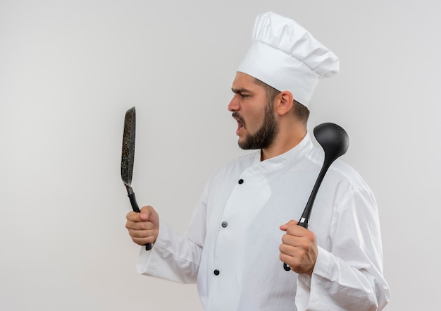 Cocinero de sexo masculino joven enojado en uniforme del cocinero que sostiene la sartén y el cucharón que miran la sartén aislada en la pared blanca