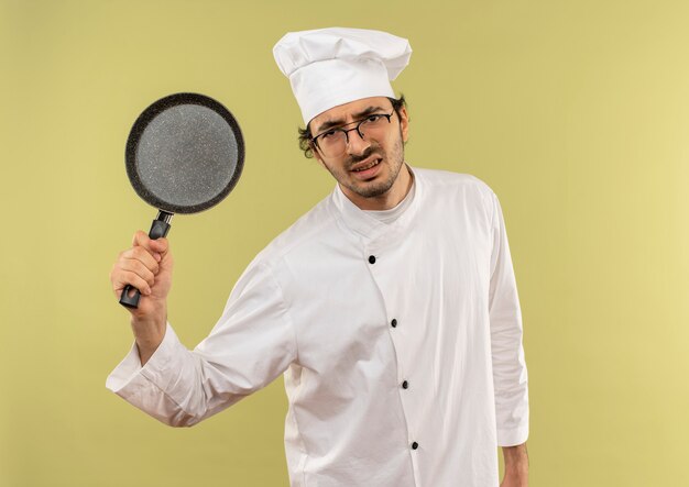 Cocinero de sexo masculino joven enojado vistiendo uniforme del cocinero y vidrios que levantan la sartén