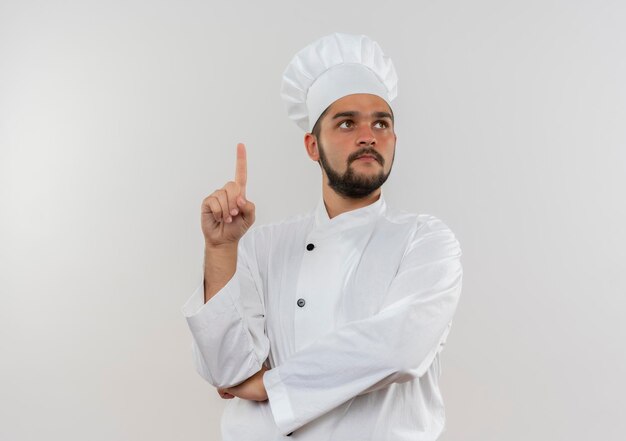 Cocinero de sexo masculino joven impresionado en uniforme del cocinero que mira al lado y que levanta el dedo aislado en la pared blanca con espacio de la copia