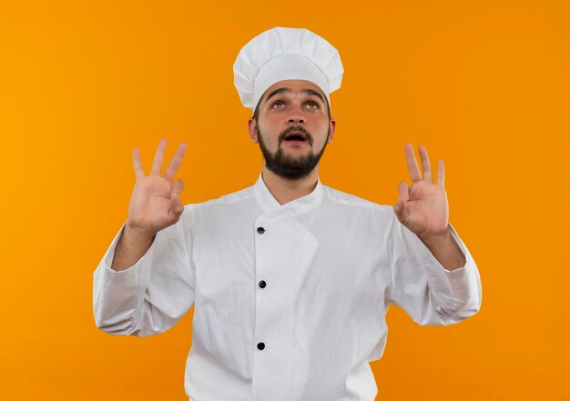 Cocinero de sexo masculino joven impresionado en uniforme del cocinero que mira hacia arriba y que hace señales bien aisladas en la pared naranja