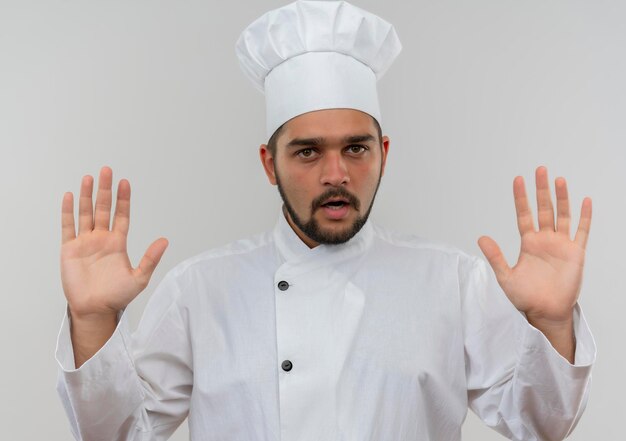 Cocinero de sexo masculino joven impresionado en uniforme del cocinero que muestra las manos vacías aisladas en la pared blanca