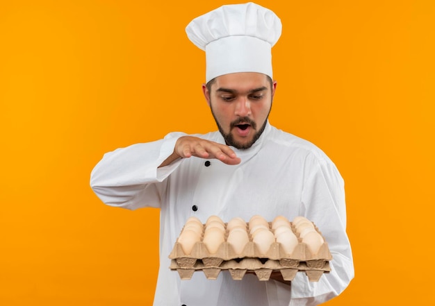 Cocinero de sexo masculino joven impresionado en uniforme del cocinero que sostiene y que mira el cartón de huevos y que mantiene la mano en el aire aislado en la pared naranja