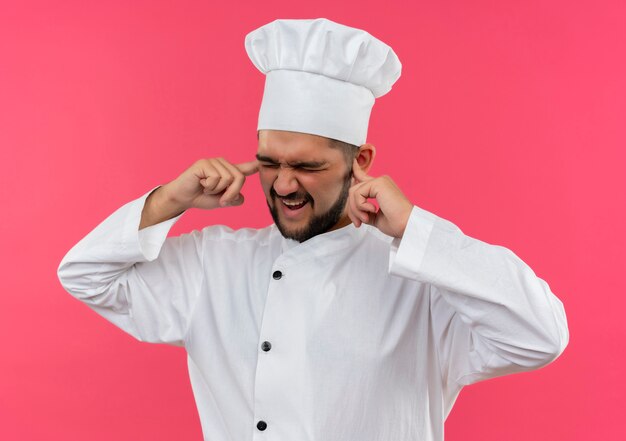 Cocinero de sexo masculino joven molesto en uniforme del cocinero que pone los dedos en los oídos con los ojos cerrados aislados en la pared rosada