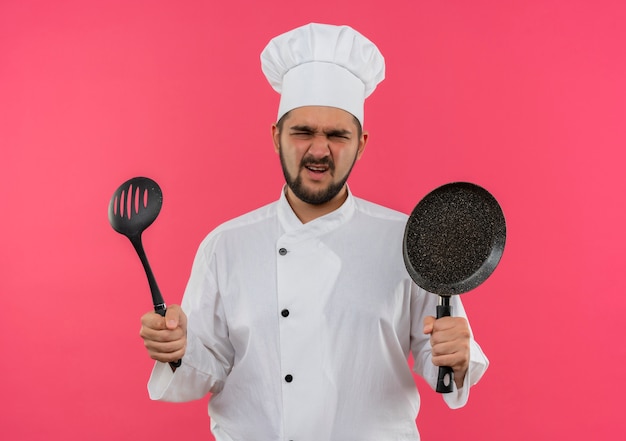 Cocinero de sexo masculino joven molesto en uniforme del cocinero que sostiene la sartén y la cuchara ranurada aislada en la pared rosada