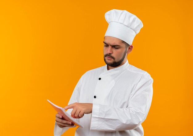 Cocinero de sexo masculino joven pensativo en uniforme del cocinero que sostiene mirando y poniendo el dedo en el bloc de notas aislado en el espacio naranja