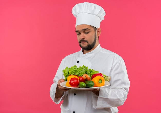Cocinero de sexo masculino joven pensativo en uniforme del cocinero que sostiene y que mira el plato de verduras aislado en el espacio rosado