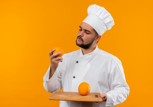 Cocinero de sexo masculino joven pensativo en uniforme del cocinero que sostiene la tabla de cortar y naranja que mira naranja aislada en el espacio anaranjado