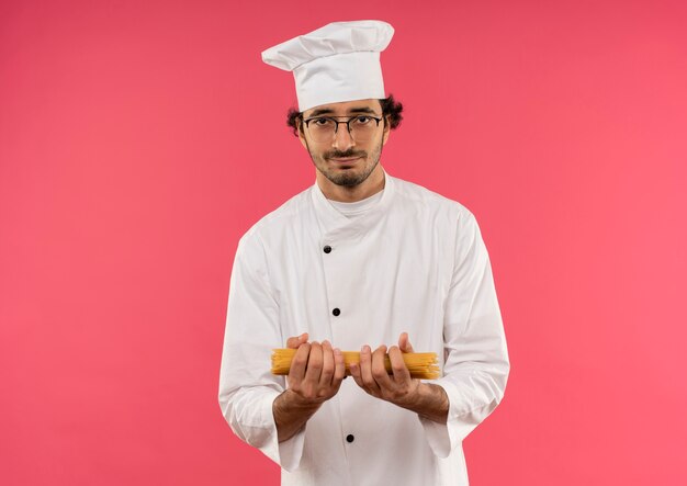 Cocinero de sexo masculino joven satisfecho con uniforme de chef y gafas sosteniendo espaguetis aislado en la pared rosa