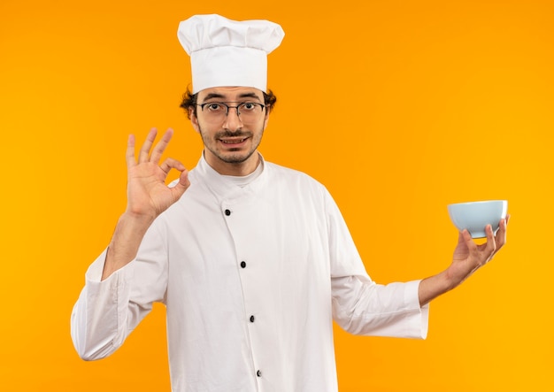 Cocinero de sexo masculino joven satisfecho con uniforme de chef y gafas sosteniendo el tazón y mostrando gesto okey aislado en la pared amarilla