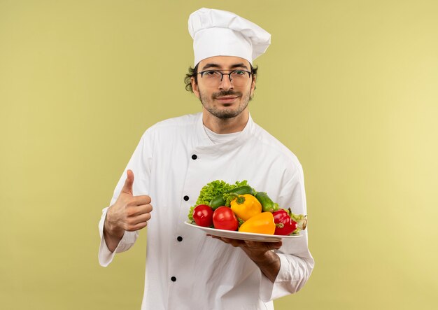 Cocinero de sexo masculino joven satisfecho con uniforme de chef y gafas sosteniendo verduras en la placa con el pulgar hacia arriba aislado en la pared verde