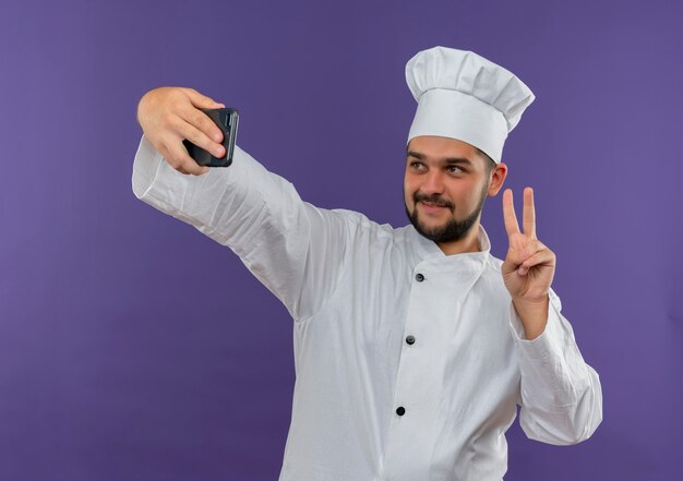 Cocinero de sexo masculino joven satisfecho en uniforme de chef haciendo el signo de la paz y tomando selfie aislado en el espacio púrpura